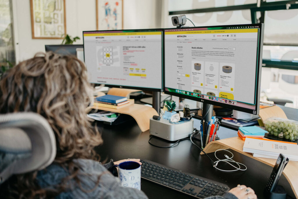 Op de voorgrond zit een medewerkster van Noeste IJver te kijken naar twee beeldschermen met daarop de Webshop van Etikon.